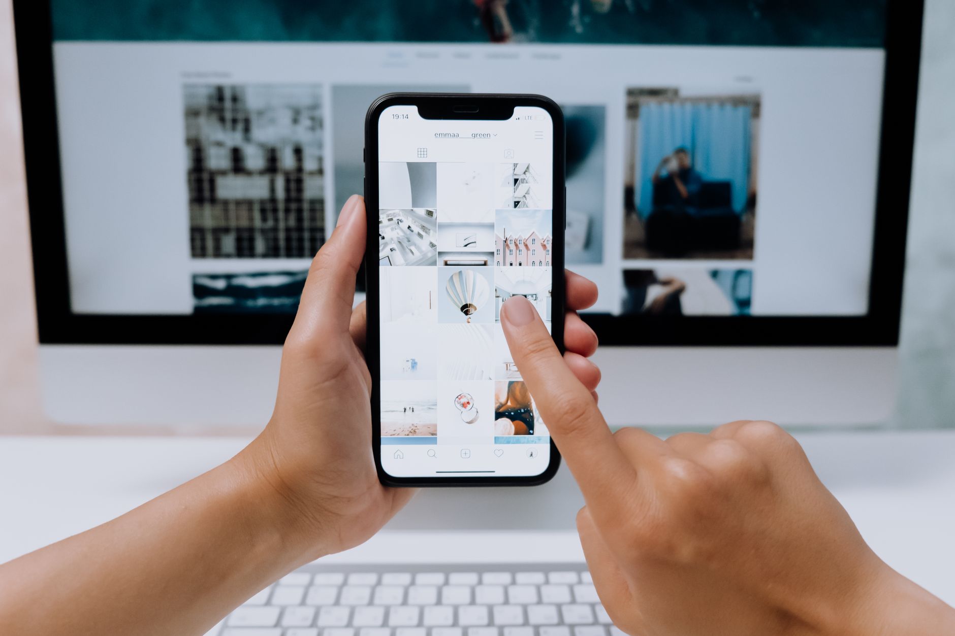 Hands scrolling a photo grid on a phone in front of a desktop monitor