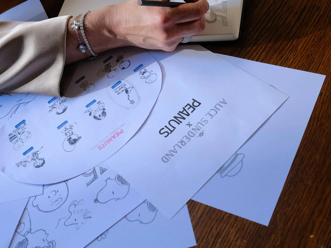 Alice Sunderland sketching Snoopy pendant designs at her desk, with the Alice Sunderland x Peanuts lockup sheet on the wood